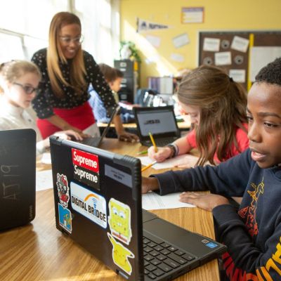 kids working on laptops with teacher in background