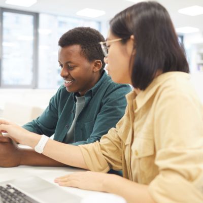 Portrait of two students studying in college library, focus on Asian young woman using laptop in foreground.