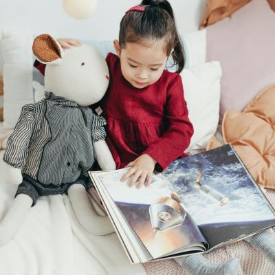 Little girl sitting on her bed with stuffed animals and reading a book.